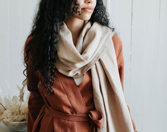 May include: A woman wearing a rust-colored linen dress with a long, beige scarf tied around her neck. The dress has a wrap style and a belt.