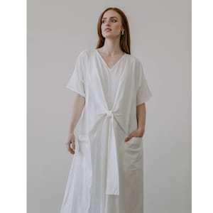 May include: A white, loose-fitting dress with a V-neck and a tie at the waist. The dress has short sleeves and two pockets.
