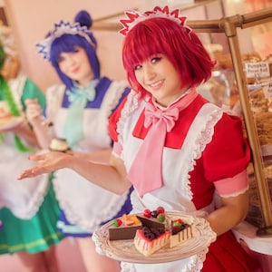 May include: Four individuals dressed in colorful maid outfits, each holding a plate with a dessert. The maid in the foreground is holding a tray with slices of cake. The background features a display case with baked goods.