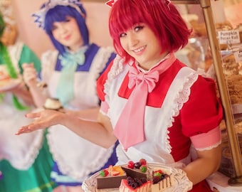 May include: Four individuals dressed in colorful maid outfits, each holding a plate with a dessert. The maid in the foreground is holding a tray with slices of cake. The background features a display case with baked goods.