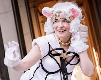 May include: A woman in a white and black dress with a fluffy white collar and matching gloves. She is wearing a white wig with pink ears and a gold necklace. She is smiling and looking to the side.