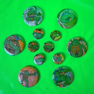 Op de afbeelding: Een verzameling Teenage Mutant Ninja Turtles-knopspelden in verschillende maten. De spelden tonen afbeeldingen van de vier schildpadden in actieposes, met groene, oranje en blauwe accenten. De achtergrond is een felgroene stof.