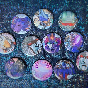 Könnte beinhalten: Zehn runde Buttons mit farbenfrohen Illustrationen von Transformers-Charakteren. Die Buttons zeigen Robotergesichter und -designs in Blau-, Lila-, Silber- und Rottönen. Sie sind auf einer schimmernden, dunkelblauen Oberfläche angeordnet.