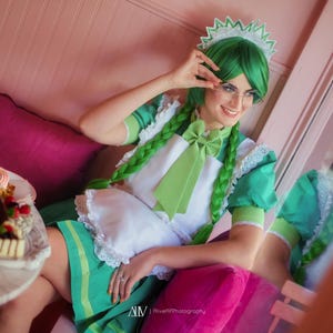 May include: A person in a green and white maid costume with green hair and a matching headpiece. The person is sitting on a pink cushion, with a plate of desserts on a table nearby. The costume includes a bow and apron.