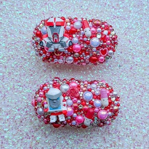 May include: Two oval-shaped hair clips decorated with a dense arrangement of red, pink, and white beads, pearls, and rhinestones. Each clip features a small robot figurine and two miniature pill-shaped charms with smiling faces.
