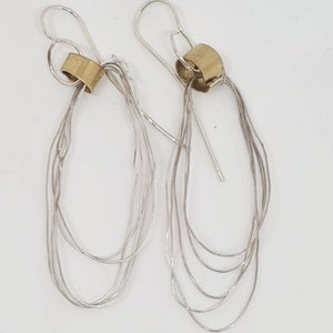 May include: A pair of silver earrings with a gold accent. The earrings are made of multiple strands of silver wire that are twisted and looped together. The gold accent is a small, round piece of metal that is attached to the top of the earrings.