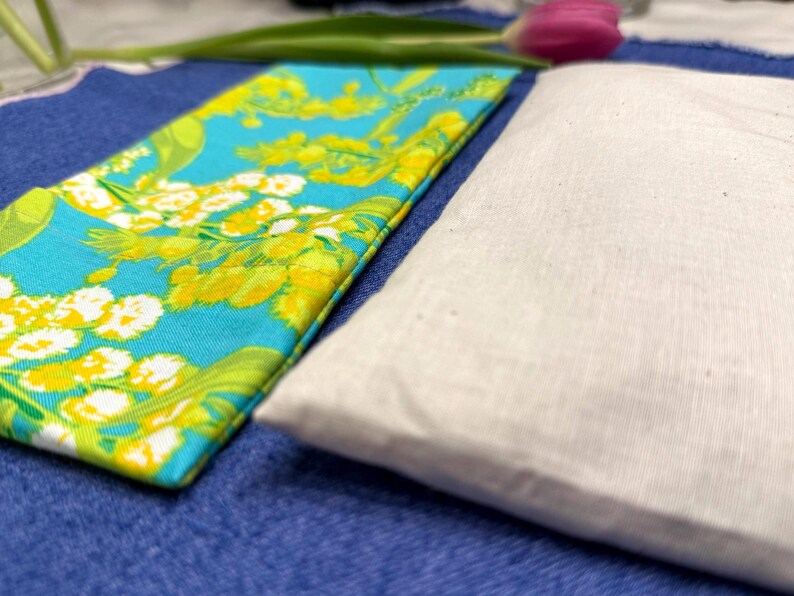 Puede incluir: Primer plano de dos art&iacute;culos de tela. Uno es una tela rectangular turquesa con un estampado floral en amarillo y blanco. El otro es una tela rectangular blanca. Ambos est&aacute;n sobre una superficie azul.