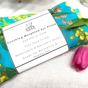 Puede incluir: Una almohada para ojos turquesa con un estampado floral en amarillo y verde. La etiqueta dice "Soothing Weighted Eye Pillow" y menciona el uso en caliente o en fr&iacute;o. Se ve un tulip&aacute;n rosa. Rellena de semillas de lino.