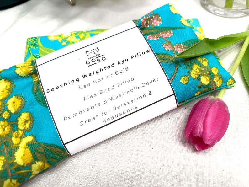 Puede incluir: Una almohada para ojos turquesa con un estampado floral en amarillo y verde. La etiqueta dice "Soothing Weighted Eye Pillow" y menciona el uso en caliente o en fr&iacute;o. Se ve un tulip&aacute;n rosa. Rellena de semillas de lino.