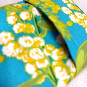 Puede incluir: Un art&iacute;culo de tela doblada con un vibrante estampado floral. El dise&ntilde;o presenta flores blancas y amarillas con hojas verdes sobre un fondo turquesa brillante. La tela est&aacute; doblada, revelando el patr&oacute;n.