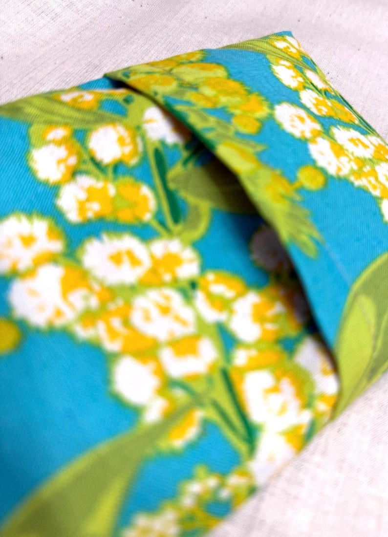 Puede incluir: Un art&iacute;culo de tela doblada con un vibrante estampado floral. El dise&ntilde;o presenta flores blancas y amarillas con hojas verdes sobre un fondo turquesa brillante. La tela est&aacute; doblada, revelando el patr&oacute;n.