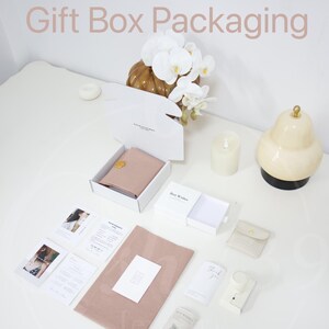 May include: An overhead shot of gift box packaging with a white box containing a blush-colored item, cards, and a small pouch. The words "Gift Box Packaging" are displayed in a light brown font. Decorative elements include a candle, a pear-shaped lamp, and a floral arrangement.