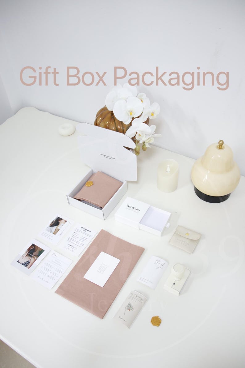 May include: An overhead shot of gift box packaging with a white box containing a blush-colored item, cards, and a small pouch. The words "Gift Box Packaging" are displayed in a light brown font. Decorative elements include a candle, a pear-shaped lamp, and a floral arrangement.