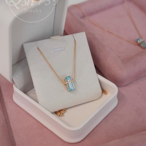 May include: A gold necklace with a rectangular, light blue gemstone pendant is displayed in a white jewelry box. The box is open, revealing the necklace on a white card. The background is a soft pink, with another necklace visible.