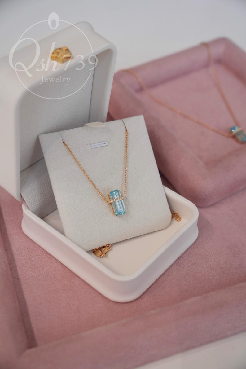 May include: A gold necklace with a rectangular, light blue gemstone pendant is displayed in a white jewelry box. The box is open, revealing the necklace on a white card. The background is a soft pink, with another necklace visible.