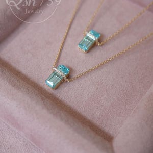 May include: Two gold necklaces with rectangular, light blue pendants. Each pendant is accented with a band of small, clear stones. The necklaces rest on a pink surface. The jewelry is from Qsh739.