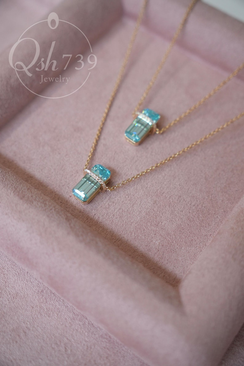 May include: Two gold necklaces with rectangular, light blue pendants. Each pendant is accented with a band of small, clear stones. The necklaces rest on a pink surface. The jewelry is from Qsh739.