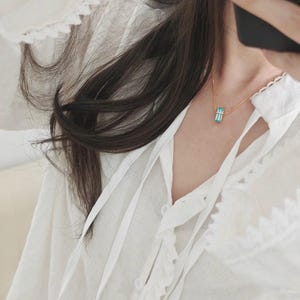May include: A gold-colored necklace with a rectangular, light blue pendant. The pendant is suspended from a delicate chain. The necklace is worn over a white blouse with a ruffled collar and tie detail.