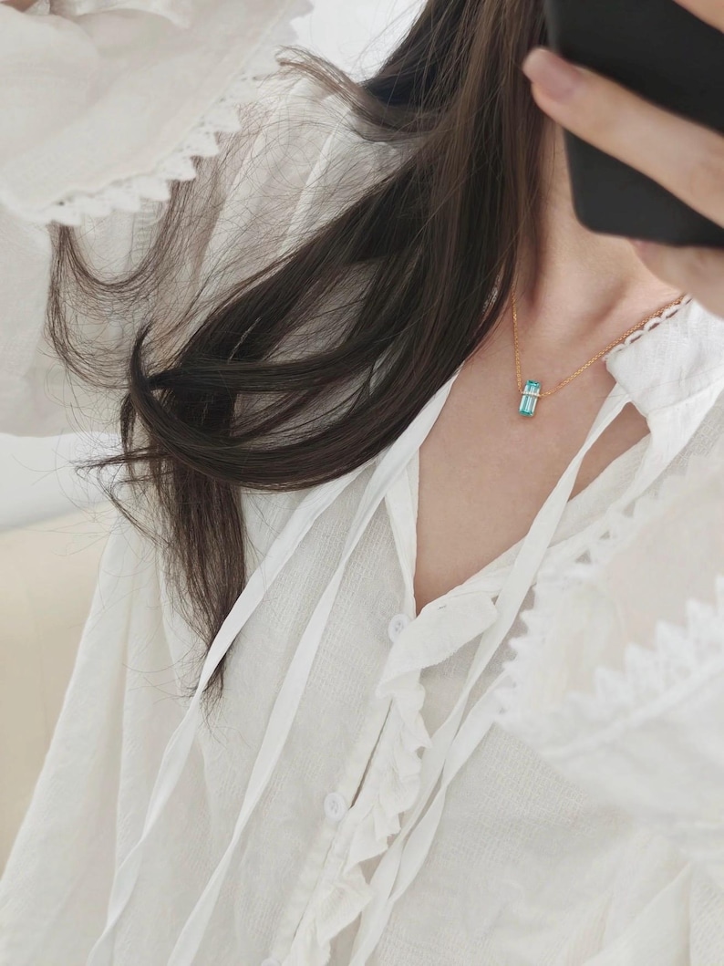 May include: A gold-colored necklace with a rectangular, light blue pendant. The pendant is suspended from a delicate chain. The necklace is worn over a white blouse with a ruffled collar and tie detail.