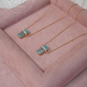 May include: Two gold-toned necklaces with rectangular, light blue pendants. Each pendant is accented with a band of small, clear stones. The necklaces rest on a pink, velvet-like surface. The jewelry is from Qsh739.