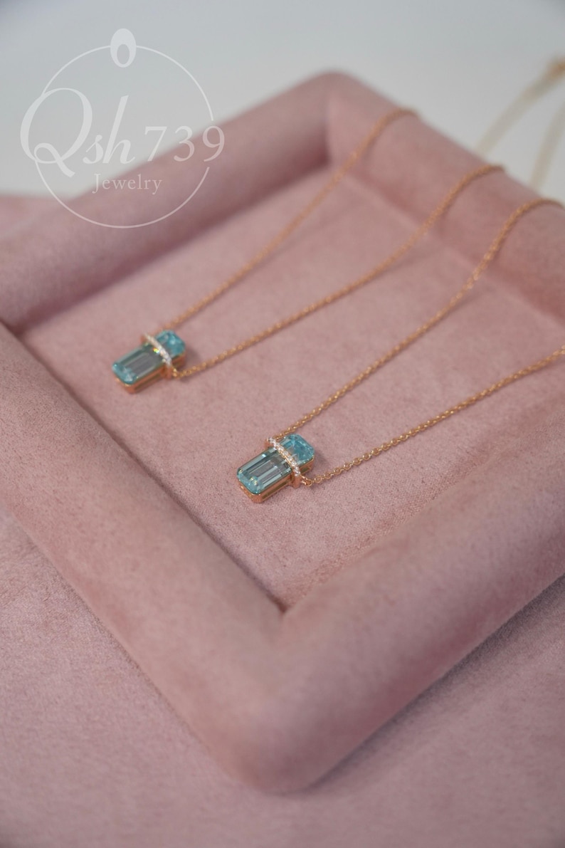 May include: Two gold-toned necklaces with rectangular, light blue pendants. Each pendant is accented with a band of small, clear stones. The necklaces rest on a pink, velvet-like surface. The jewelry is from Qsh739.