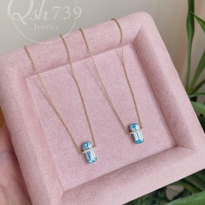 May include: Two gold necklaces with rectangular, light blue pendants. Each pendant is accented with a band of small, clear stones. The necklaces are displayed on a pink tray, with the jewelry brand name "Qsh739 Jewelry" in the background.