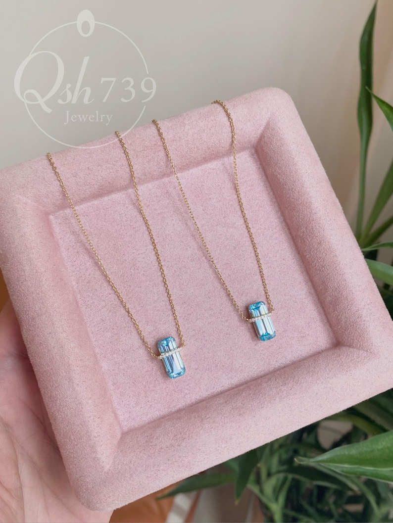 May include: Two gold necklaces with rectangular, light blue pendants. Each pendant is accented with a band of small, clear stones. The necklaces are displayed on a pink tray, with the jewelry brand name "Qsh739 Jewelry" in the background.