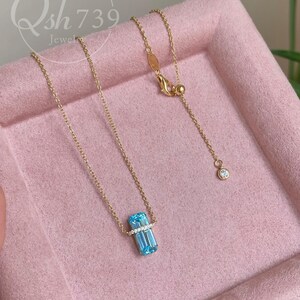 May include: A delicate gold necklace featuring a rectangular, light blue gemstone pendant with a row of small, clear stones. The necklace has a fine chain and a small, round accent stone near the clasp. The pendant is approximately 0.5 inches long.