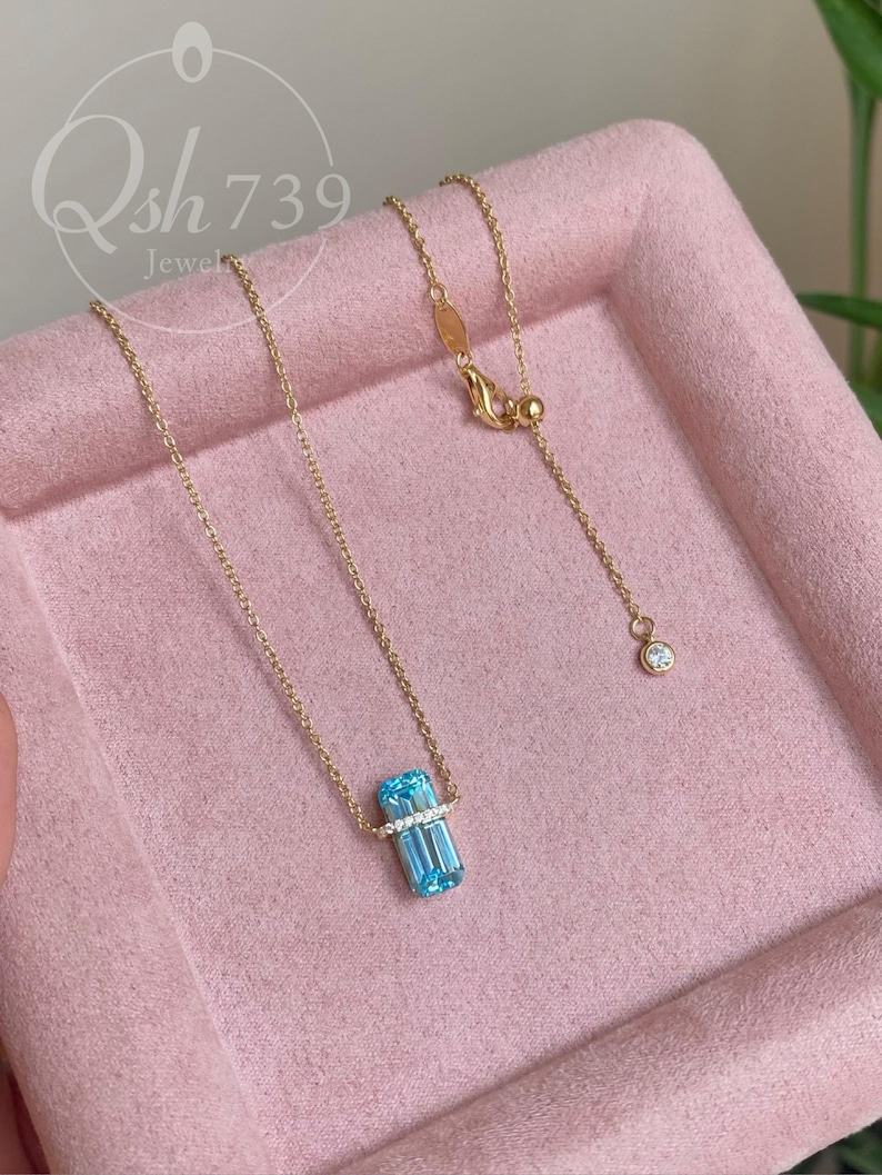 May include: A delicate gold necklace featuring a rectangular, light blue gemstone pendant with a row of small, clear stones. The necklace has a fine chain and a small, round accent stone near the clasp. The pendant is approximately 0.5 inches long.