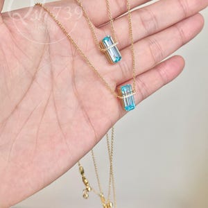 May include: A delicate gold necklace featuring two strands. Each strand has a rectangular, light blue gemstone pendant with a halo of small, clear stones. The necklace is displayed on a hand.