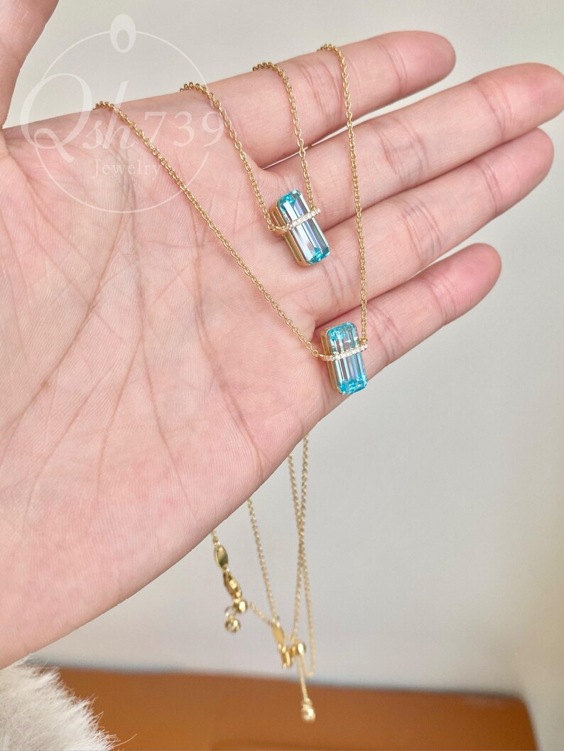 May include: A delicate gold necklace featuring two strands. Each strand has a rectangular, light blue gemstone pendant with a halo of small, clear stones. The necklace is displayed on a hand.