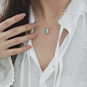May include: A gold necklace featuring a rectangular, light blue gemstone pendant. The pendant is suspended from a delicate gold chain. The necklace is worn against a white blouse.