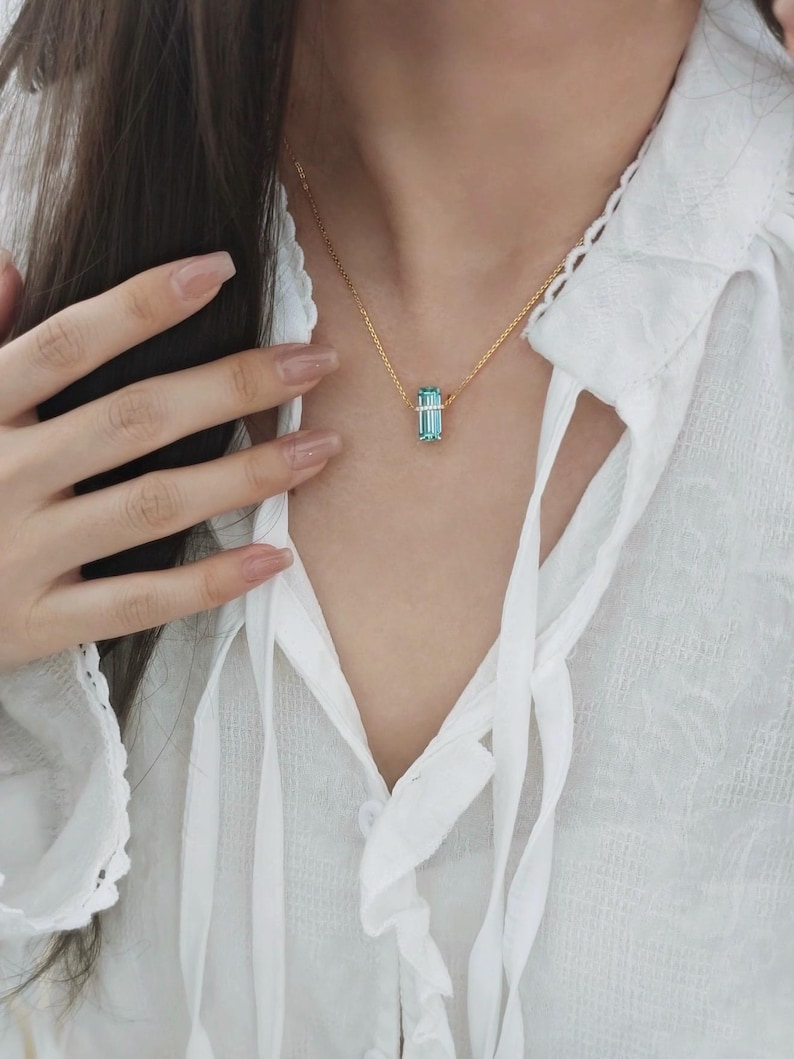 May include: A gold necklace featuring a rectangular, light blue gemstone pendant. The pendant is suspended from a delicate gold chain. The necklace is worn against a white blouse.