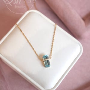 May include: A gold chain necklace with a rectangular, light blue gemstone pendant. The pendant is set with small, clear stones. The necklace is displayed on a white surface against a pink background. The text "Qsh 739 Jewelry" is in the upper left corner.