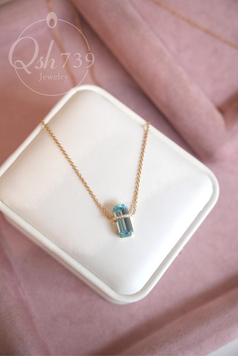 May include: A gold chain necklace with a rectangular, light blue gemstone pendant. The pendant is set with small, clear stones. The necklace is displayed on a white surface against a pink background. The text "Qsh 739 Jewelry" is in the upper left corner.
