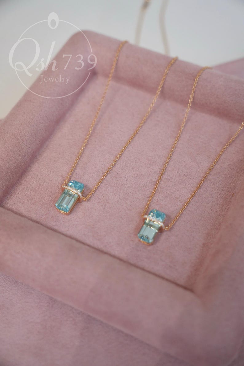 May include: Two gold necklaces with rectangular, light blue gemstone pendants. Each pendant is accented with small, clear stones. The necklaces are displayed on a pink surface. The jewelry is from Qsh739 jewelry.