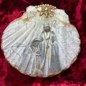 Può includere: Conchiglia dipinta a mano raffigurante una scena religiosa con due figure in abiti, una stella e bordi dorati. Un abbellimento decorativo con gioielli è attaccato alla parte superiore della conchiglia. Lo sfondo è in velluto rosso scuro.