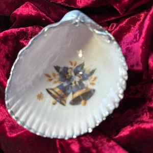 May include: A decorative seashell dish with a silver-colored trim. The interior features a design of blue bells with gold accents and leaves. The dish is set against a deep red velvet background.