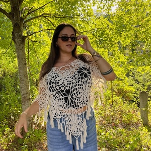 Puede incluir: Top de crochet crema de tejido abierto con diseño floral y flecos. El top se lleva sobre una prenda negra y se combina con vaqueros azules. El fondo presenta un exuberante follaje verde y luz solar.