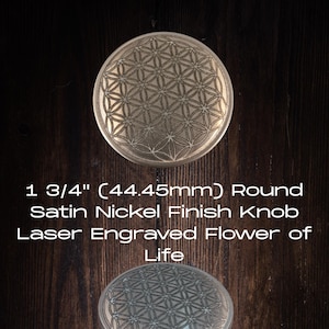 Può includere: Due pomelli rotondi in nichel satinato con un design a Fiore della Vita inciso al laser. Ogni pomello misura 44,45 mm di diametro. I pomelli sono esposti su uno sfondo di venature del legno scuro.