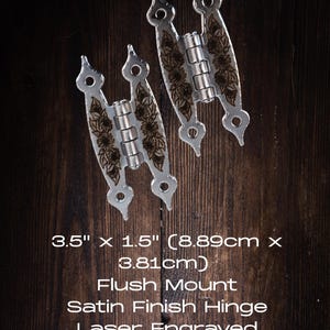 Może przedstawiać: Dwa zawiasy do montażu równo z powierzchnią o satynowym wykończeniu. Każdy zawias ma wymiary 8,89 cm x 3,81 cm i posiada grawerowane laserowo słoneczniki. Tekst na zdjęciu brzmi: "Flush Mount, Satin Finish Hinge, Laser Engraved Sunflowers."