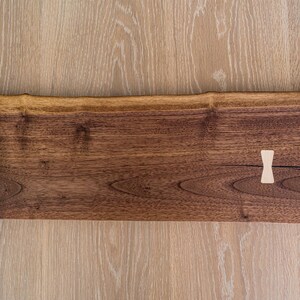 May include: A long, rectangular wooden serving tray with a natural wood finish. The tray has a live edge and two small wooden pegs on the surface.