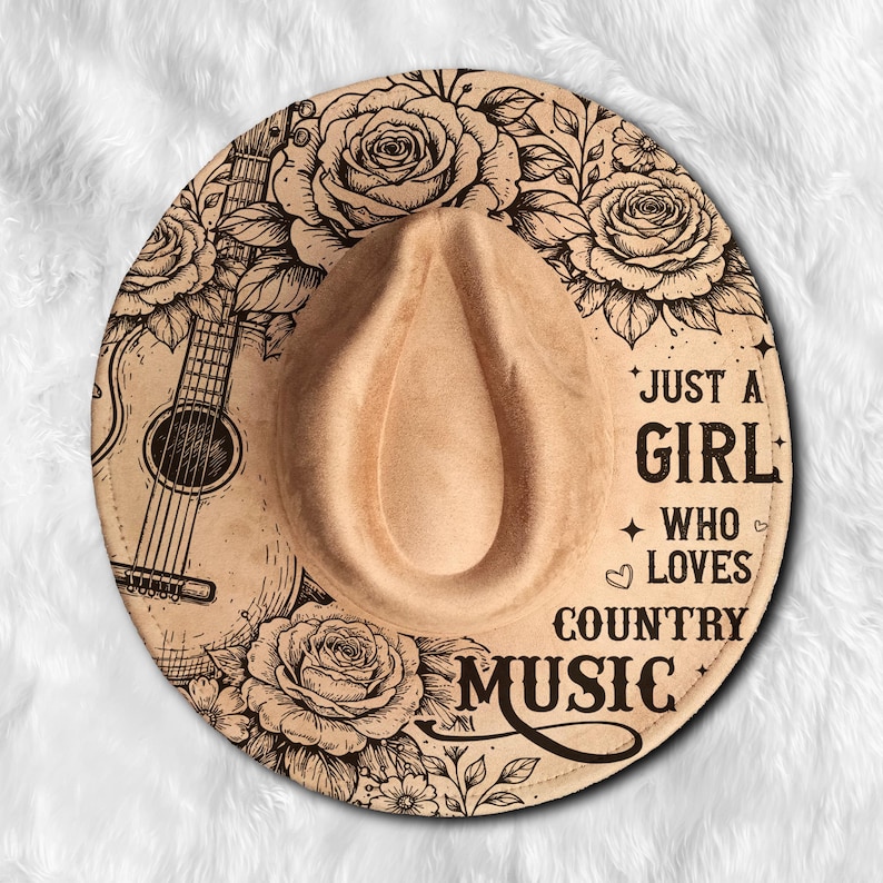 May include: Tan fedora-style hat with a floral and guitar design. The hat features the text "JUST A GIRL WHO LOVES COUNTRY MUSIC" in black. The hat has a teardrop crown and a wide brim.