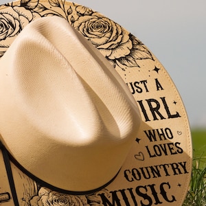 May include: A beige cowboy hat with a curved brim. The hat's band features the text "Just a Girl Who Loves Country Music" in black, along with rose illustrations. The hat is set against a blurred green background.