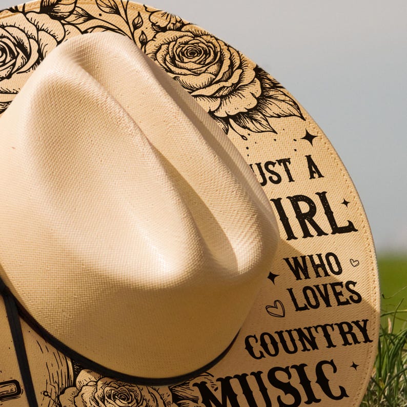 May include: A beige cowboy hat with a curved brim. The hat's band features the text "Just a Girl Who Loves Country Music" in black, along with rose illustrations. The hat is set against a blurred green background.
