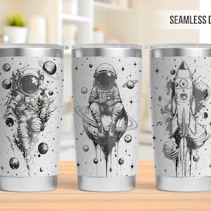 May include: Three stainless steel tumblers with a white background and black space-themed designs. The designs include an astronaut, a rocket ship, and a planet. The tumblers are all the same size and shape.