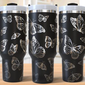 Puede incluir: Taza de viaje de acero inoxidable negro con un diseño de mariposa plateada. La taza tiene un asa y una tapa. "Seamless Design" está impreso en la esquina inferior derecha de la imagen.