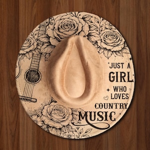 May include: A tan cowboy hat with a decorative floral and guitar design. The hat features the text "Just a Girl Who Loves Country Music" in black lettering. The hat is set against a wooden background.