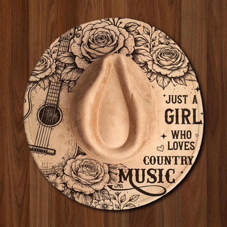 May include: A tan cowboy hat with a decorative floral and guitar design. The hat features the text "Just a Girl Who Loves Country Music" in black lettering. The hat is set against a wooden background.