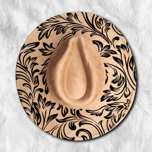 May include: A tan fedora-style hat with a wide brim and a teardrop-shaped crown. The brim is decorated with a black floral pattern. The hat is made of a soft, suede-like material.